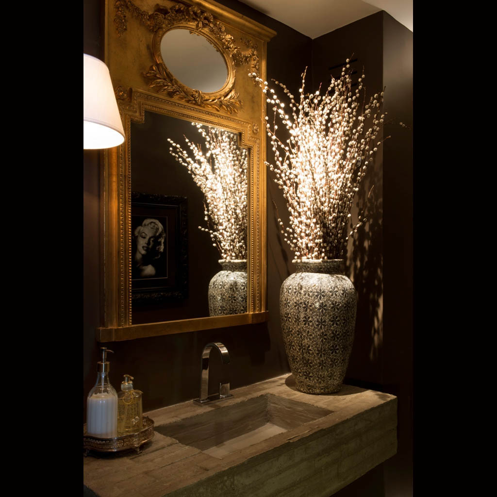 No lavabo, a bancada contemporânea em concreto moldada no próprio local contrasta com o espelho clássico dourado